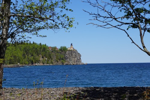 A lighthouse perched on a rocky cliff, surrounded by trees and overlooking a calm blue lake under a clear sky.