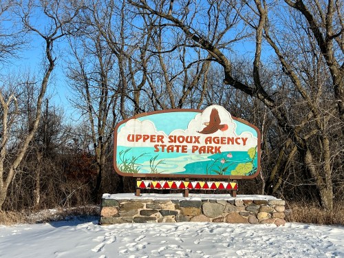 Sign for Upper Sioux Agency State Park, featuring colorful artwork and surrounded by trees and snow.