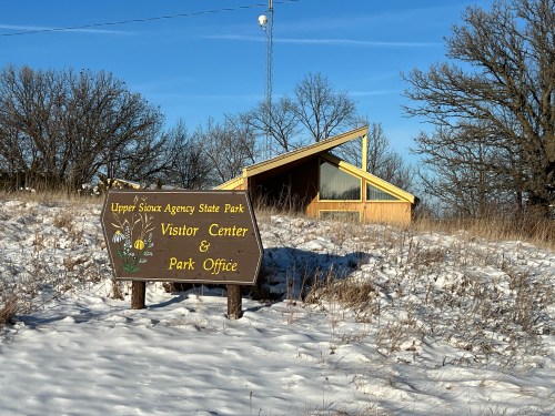 Sign for Upper Sioux Agency State Park Visitor Center and Park Office, with a modern building and snow-covered ground.