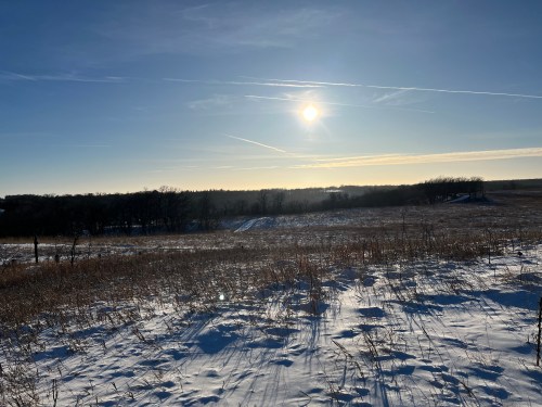 A snowy landscape under a clear blue sky, with the sun shining brightly and trees in the distance.
