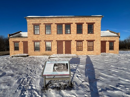 Historic brick building with multiple windows and doors, surrounded by snow, featuring an informational sign in front.