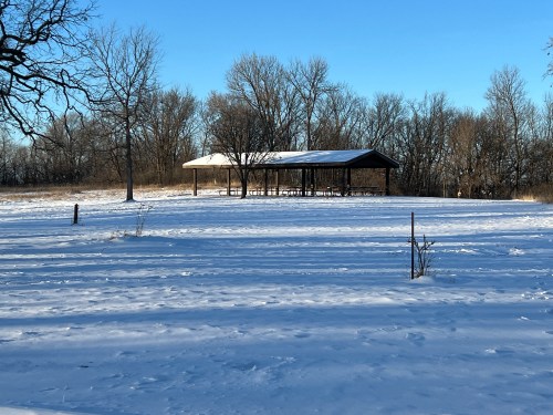 A snow-covered field with a pavilion in the background, surrounded by bare trees under a clear blue sky.