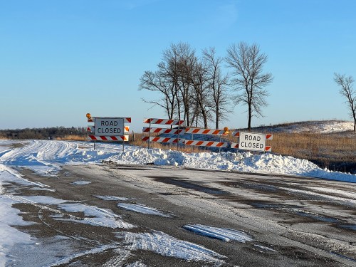 A snow-covered road with barriers and signs indicating "Road Closed" in a rural area. Trees are visible in the background.
