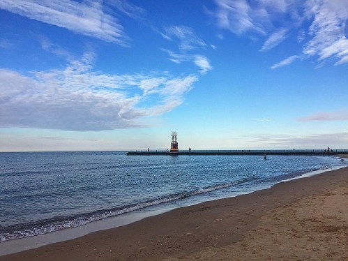 A serene beach scene with gentle waves, a lighthouse on a pier, and a clear blue sky with scattered clouds.