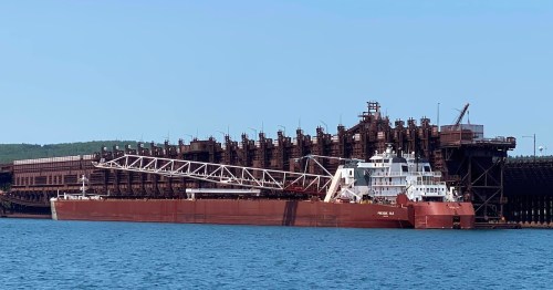A large cargo ship docked at a port, with industrial structures in the background and calm water in the foreground.