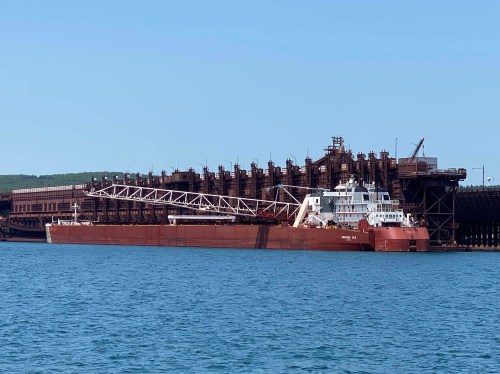 A large cargo ship docked at a port, with industrial structures and a clear blue sky in the background.
