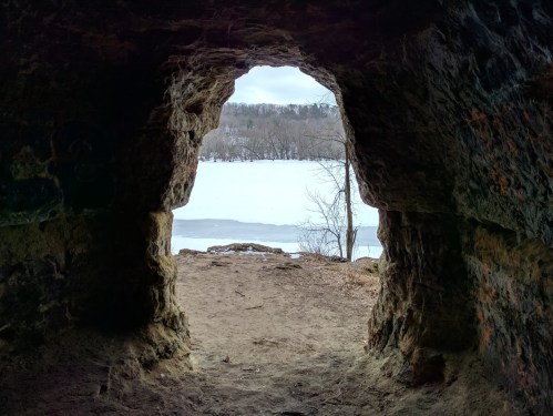 View from a cave opening, looking out at a snowy landscape and a frozen lake surrounded by trees.