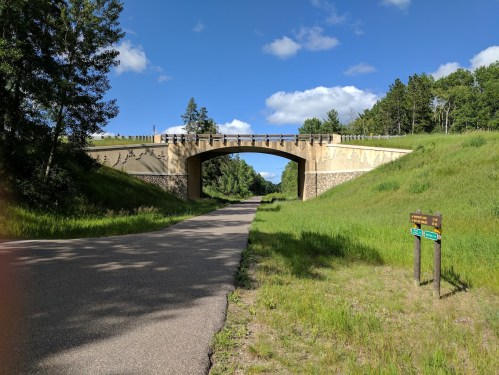 A scenic view of a bridge over a road, surrounded by lush green grass and trees under a blue sky with clouds.