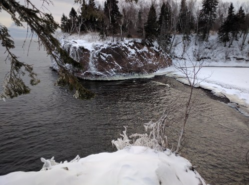 A snowy shoreline with a rocky outcrop and trees, overlooking a calm body of water.