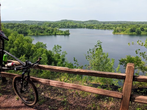 A bike rests against a wooden fence overlooking a serene lake surrounded by lush greenery and rolling hills.