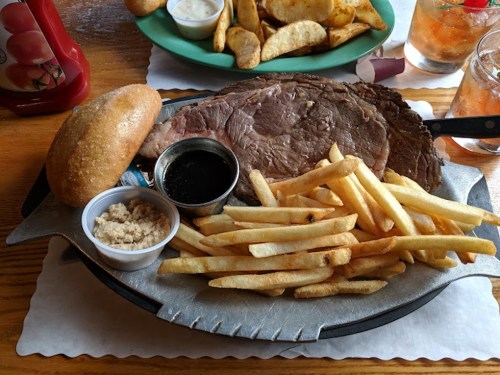 A sizzling platter with a large cut of prime rib, fries, a roll, and dipping sauce, served in a casual dining setting.