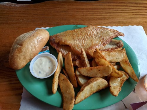 A green plate with fried fish, seasoned potato wedges, a roll, and a small cup of dipping sauce.