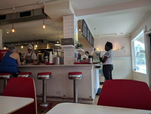 A cozy café scene with a counter, bar stools, and a woman standing near the entrance while staff prepare food.
