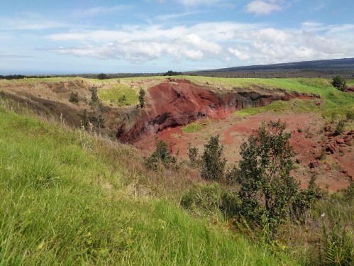 A vibrant landscape featuring a red-hued cliff surrounded by green grass and trees under a blue sky with clouds.