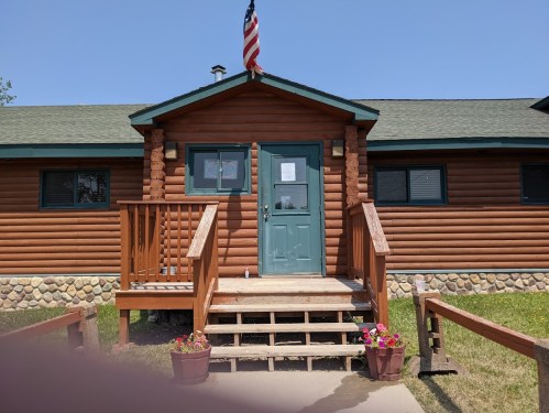 Log cabin-style building with a green door, wooden steps, and flower pots, featuring an American flag on the roof.
