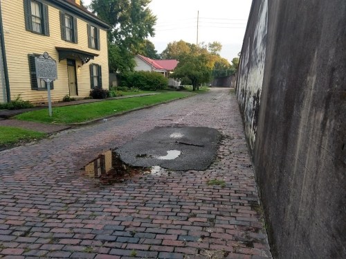 A cobblestone street with a large puddle and a historic house on one side, bordered by a tall wall.