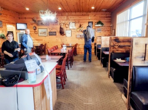 A rustic restaurant interior with wooden decor, empty tables, and a cashier area; a staff member and two patrons are present.