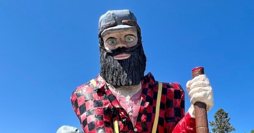 A large, colorful statue of a lumberjack with a beard, wearing a red and black checkered shirt, against a blue sky.