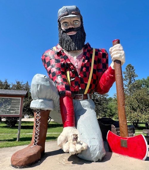 A large statue of a lumberjack in a plaid shirt, kneeling with an axe, surrounded by trees and blue sky.