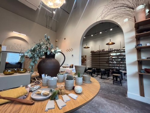 A cozy shop interior featuring a wooden table with decor items, plants, and shelves filled with pottery and goods.