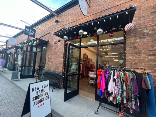 A storefront decorated with holiday lights and ornaments, featuring a clothing rack outside and a sign promoting shopping.