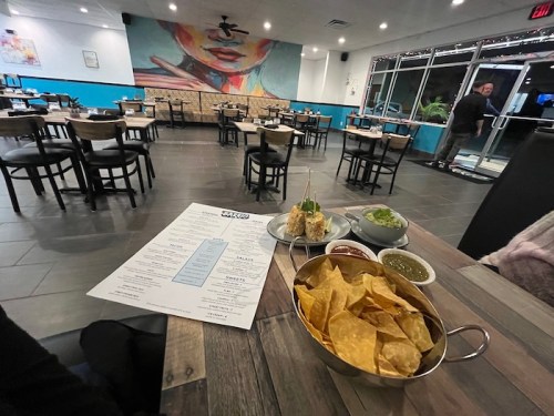A table with a menu, a bowl of tortilla chips, and three dips in a vibrant restaurant with colorful wall art.