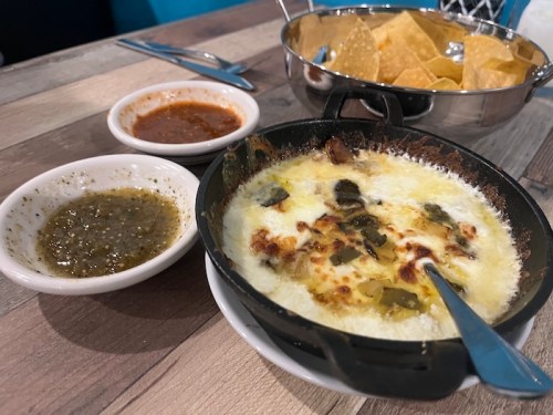 A sizzling dish with melted cheese and toppings, served with two sauces and a side of tortilla chips.