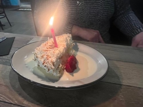 A slice of cake with a candle and a raspberry on a plate, set on a wooden table.