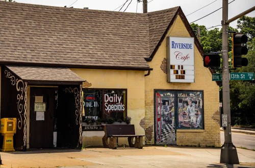 A quaint café with a sign reading "Riverside Café," featuring a mural and a bench outside, located at a street corner.