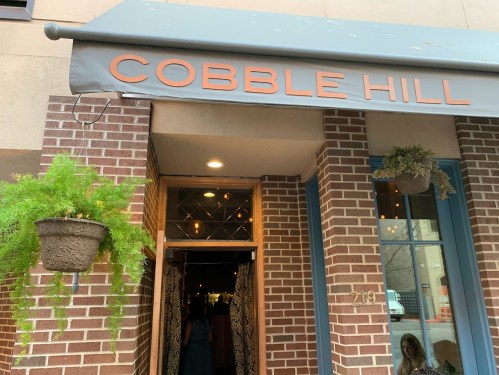 Entrance of Cobble Hill restaurant with a gray awning and hanging plants, featuring a brick facade.