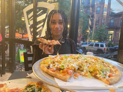 A person with braided hair holds a slice of pizza, smiling in a restaurant with a view of the street outside.