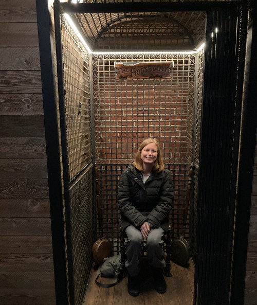 A woman sits inside a vintage-style elevator with a metal grate, smiling in a cozy, dimly lit space.