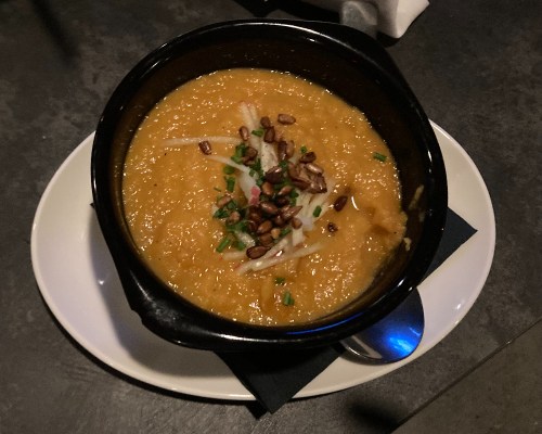 A black bowl filled with creamy orange soup, topped with green onions and toasted seeds, on a white plate.