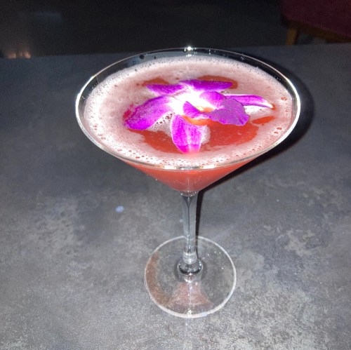A vibrant cocktail in a martini glass, topped with a purple flower, resting on a dark surface.