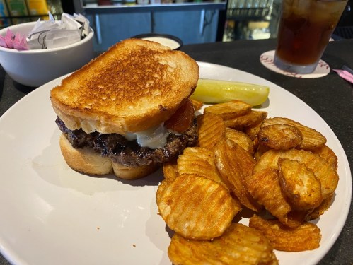 A toasted sandwich with a beef patty, tomato, and mayo, served with crispy potato chips and a pickle on the side.
