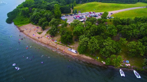 Aerial view of a beach area with people, trees, and parked cars near a lake on a sunny day.