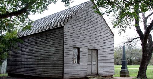 A small, weathered wooden building with a gabled roof, surrounded by trees and grass.