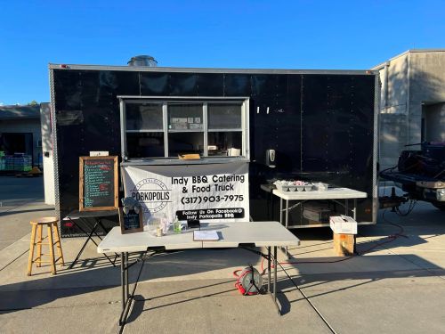 A food truck with a black exterior, featuring a service window and a table displaying a menu for BBQ catering.