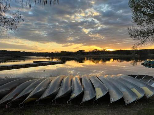 Sunset over a calm lake with stacked canoes in the foreground and a cloudy sky reflecting on the water.