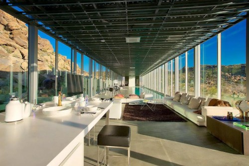 Modern interior of a glass-walled home with a sleek kitchen and living area, surrounded by rocky desert landscape.
