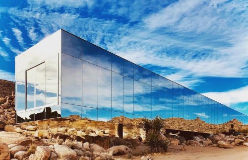 A modern, mirrored building reflects the sky and surrounding rocky landscape under a bright blue sky.