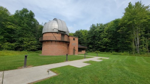 A brick observatory with a dome, surrounded by green trees and a grassy area, under a cloudy sky.