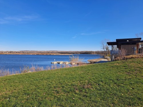 A serene lake view with a grassy shore, a dock, and a modern building under a clear blue sky.