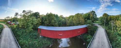 A red covered bridge spans a calm stream, surrounded by lush greenery and a clear blue sky.