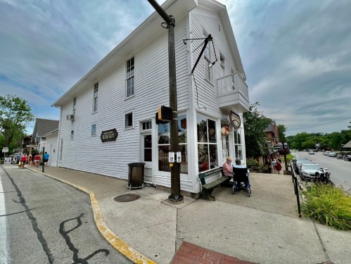 A white building with large windows, a bench outside, and a street view of a small town on a cloudy day.