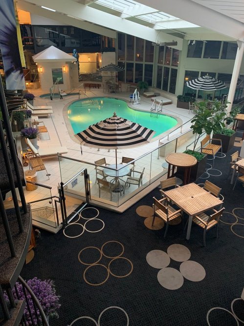 Indoor pool area with a striped umbrella, lounge chairs, and tables surrounded by greenery and patterned flooring.