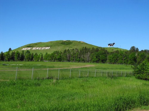 A green hill with "NEW SALEM" written on it, featuring a large cow statue on top, surrounded by trees and grass.
