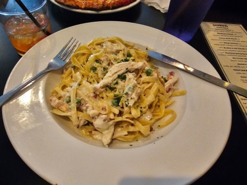 A plate of fettuccine pasta with creamy sauce, chicken, peas, and herbs, served with a drink and menu in the background.