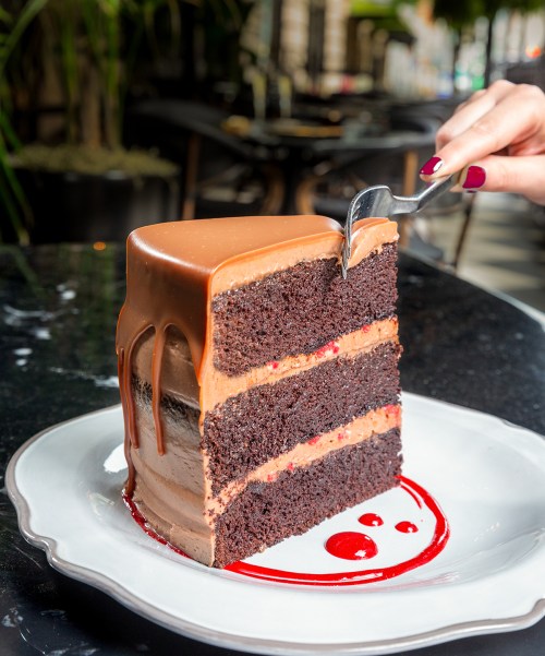 A slice of rich chocolate cake with layers and a glossy chocolate ganache, served on a white plate with a red sauce.