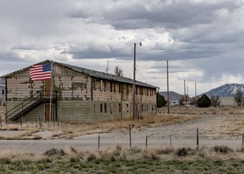 A weathered building with an American flag, set against a cloudy sky and sparse landscape.